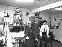 Three generations of John M Kennas barbers, Westport, February 1982.. - Lyons0014108.jpg  Three generations of John M Kennas barbers, Westport, February 1982.  At left is John Mc Kenna senior long established barber shop on Bridge St. Westport where John and his family also lived. Standing is his son Sean who took over the business when his father retired in his late 80's and next to Sean is his young son John who now runs the Gents Barber Shop in the new location  Market Lane, Bridge St. There is a new change in tradition which his grabdfather would never have dreamt of. John employs three lady barbers. Note the clock on the wall; at left St MartinDeporres black Saint and at right the infant of prague. These were possibly placed by John's senior wife Mrs Kathleen Mc Kenna. Also note each side of the wallcabinet are the rows of cut throat razors which were expertly used and sharpened by John senior for close up shaves for men. Sadly this tradition did not continue for the next generations after the invention of  safety razors. : 198202 Three Generations of the Mc Kennas 1.tif, Lyons collection, Westport