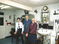 Three generations of John M Kennas barbers, Westport, February 1982.. - Lyons0014109.jpg  Three generations of John M Kennas barbers, Westport, February 1982.  At left is John Mc Kenna senior long established barber shop on Bridge St. Westport where John and his family also lived. Standing is his son Sean who took over the business when his father retired in his late 80's and next to Sean is his young son John who now runs the Gents Barber Shop in the new location  Market Lane, Bridge St. There is a new change in tradition which his grabdfather would never have dreamt of. John employs three lady barbers. Note the clock on the wall; at left St MartinDeporres black Saint and at right the infant of prague. These were possibly placed by John's senior wife Mrs Kathleen Mc Kenna. Also note each side of the wallcabinet are the rows of cut throat razors which were expertly used and sharpened by John senior for close up shaves for men. Sadly this tradition did not continue for the next generations after the invention of  safety razors. : 198202 Three Generations of the Mc Kennas 2.tif, Lyons collection, Westport